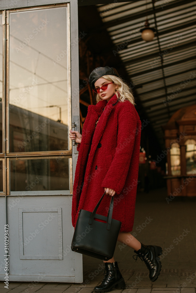 Outdoor full-length fashion portrait of young confident elegant woman wearing trendy red faux fur coat, sunglasses, beret, lace up ankle boots, holding leather bag, handbag, posing in street of city