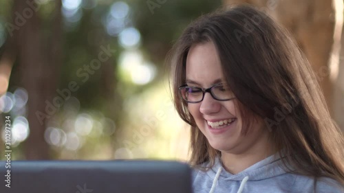 Teen girl in glasses close-up with a laptop, in the park on a bench laughs, indulges, jokes and makes funny faces.