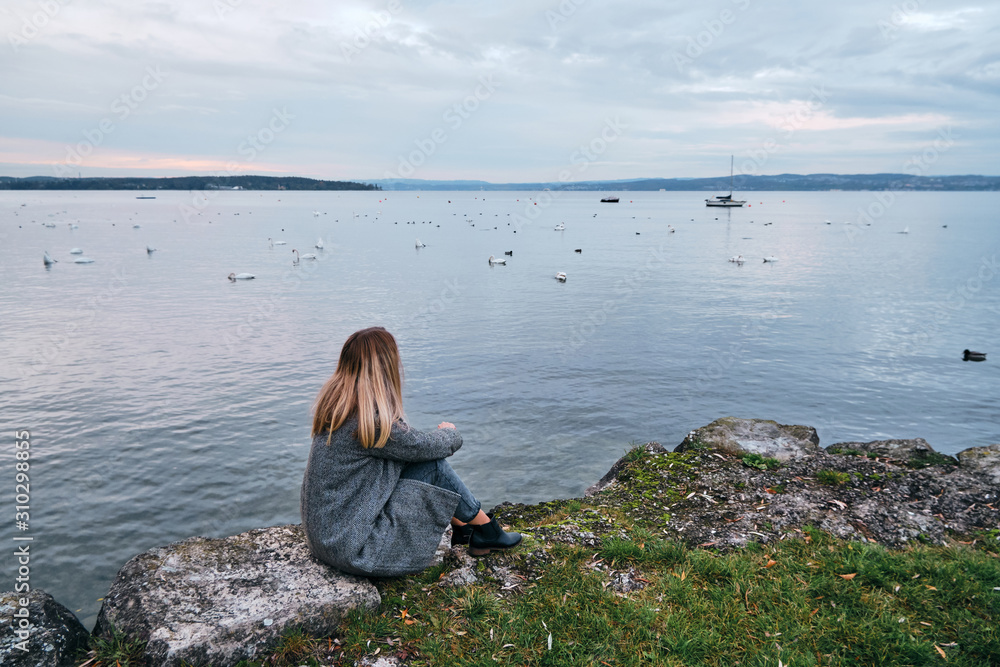 Naklejka premium Beautiful blond hair woman enjoy panorama by the Bodensee lake. Landscape in Switzerland. Happy girl in travel. Amazing scenic outdoors view. Dramatic sky. Swans on water. Adventure lifestyle, freedom