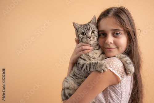 Close up portrait of the beautiful little girl with brunette loose hair the hugging her grey cat, isolated over orange background