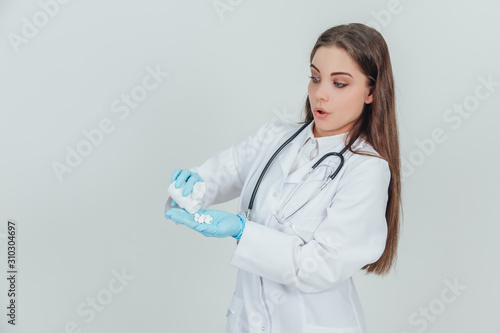 Pretty young concentrated intern girl pouring pills from bottle into her palm  dressed in disposable gloves.