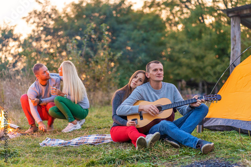 Group of happy friends with guitar, having fun outdoor, near bonfire and tourist tent. Camping fun happy family
