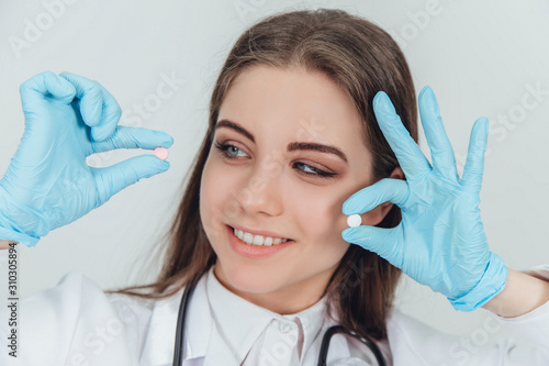 Lovely smiling young doctor holding two small white pills in her fingers near her face  looking at one of them  smiling.