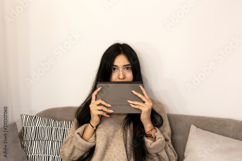 Shocked young woman with emotional face sitting on the sofa with tablet and looking surprised,