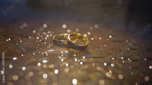 Wedding rings lying on dark surface with reflection and water. Shining with light close up macro.