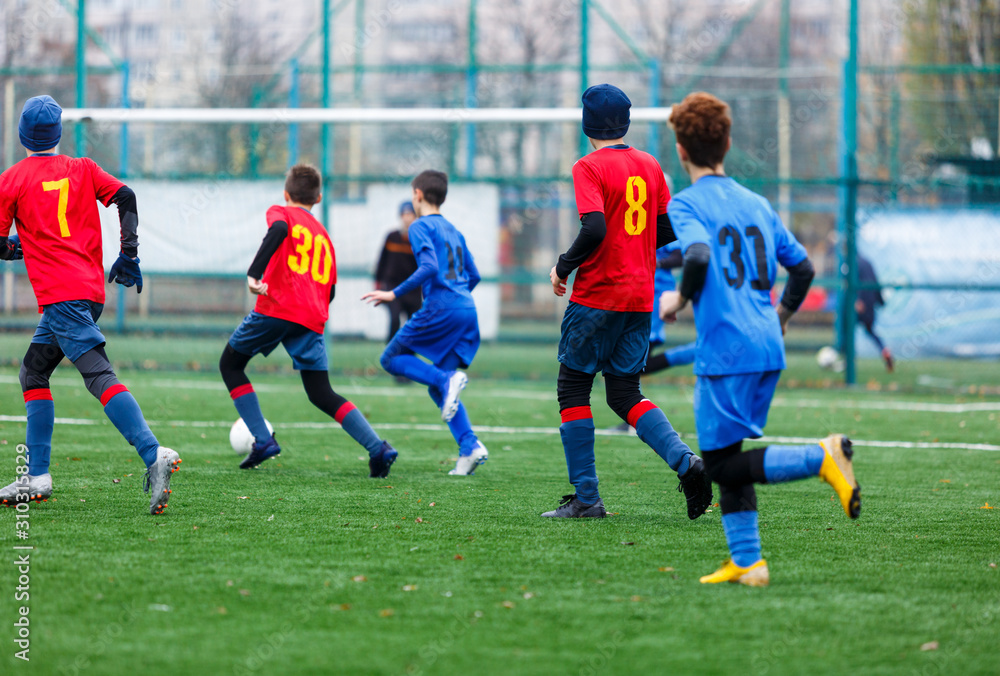 Obraz premium Boys in red and blue sportswear plays football on field, dribbles ball. Young soccer players with ball on green grass. Training, football, active lifestyle for kids concept
