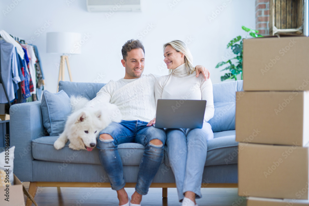 Fototapeta premium Young beautiful couple with dog sitting on the sofa using laptop at new home around cardboard boxes
