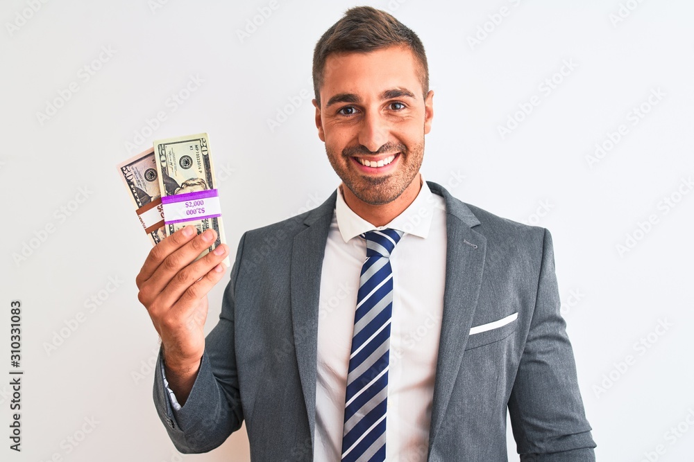 Young handsome business man holding bunch of dollars banknotes over isolated background with a happy face standing and smiling with a confident smile showing teeth Young handsome business man holding bunch of dollars banknotes over isolated background with a happy face standing and smiling with a confident smile showing teeth