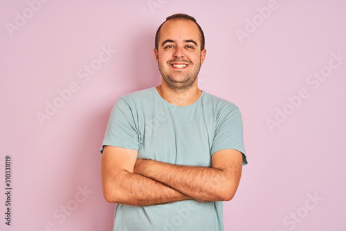 Young man wearing blue casual t-shirt standing over isolated pink background happy face smiling with crossed arms looking at the camera. Positive person.