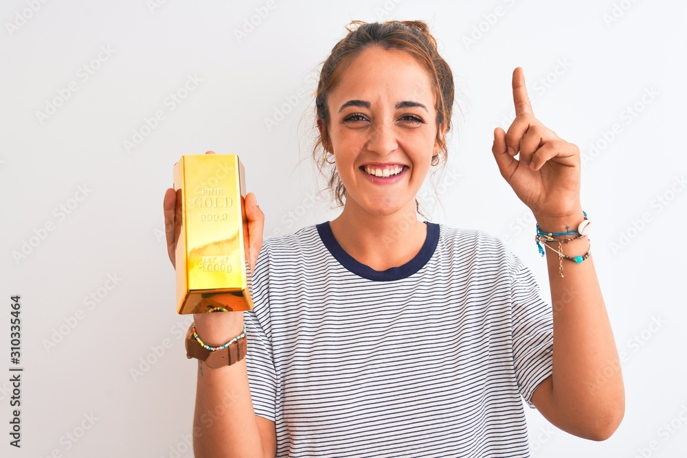 Young beautiful redhead woman holding gold ingot over isolated background surprised with an idea or question pointing finger with happy face, number one Young beautiful redhead woman holding gold ingot over isolated background surprised with an idea or question pointing finger with happy face, number one