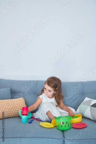 Adorable blonde toddler siting on the sofa cooking with food and cutlery toys at home