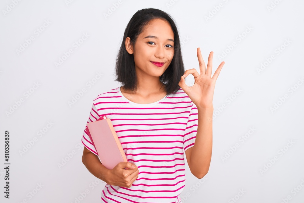 Young beautiful chinese woman holding books standing over isolated white background doing ok sign with fingers, excellent symbol Young beautiful chinese woman holding books standing over isolated white background doing ok sign with fingers, excellent symbol