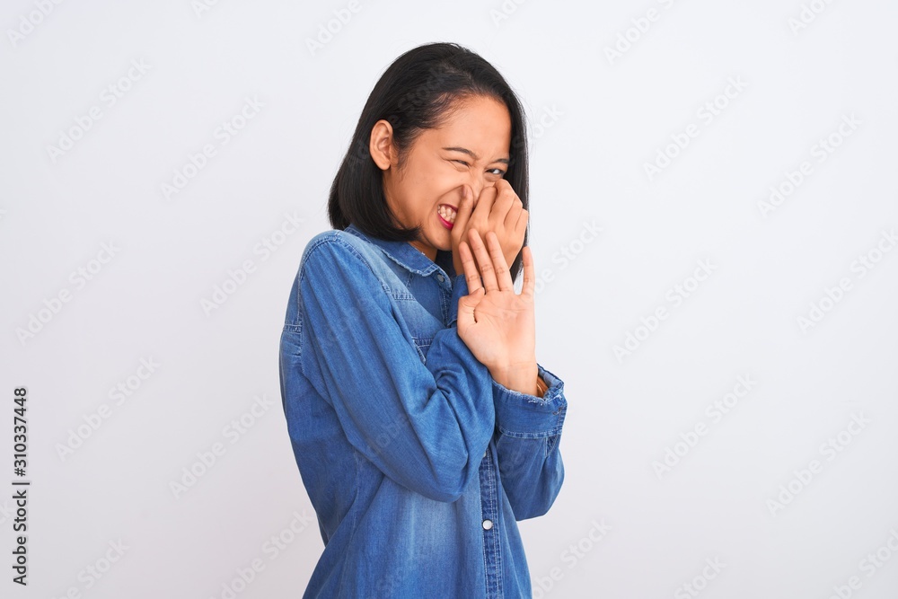Young beautiful chinese woman wearing denim shirt standing over isolated white background smelling something stinky and disgusting, intolerable smell, holding breath with fingers on nose. Bad smell Young beautiful chinese woman wearing denim shirt standing over isolated white background smelling something stinky and disgusting, intolerable smell, holding breath with fingers on nose. Bad smell