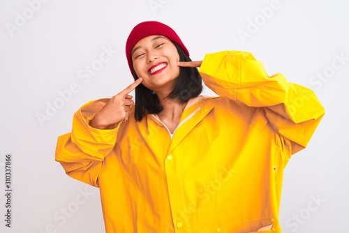 Young beautiful chinese woman wearing raincoat and wool cap over isolated white background smiling cheerful showing and pointing with fingers teeth and mouth. Dental health concept.