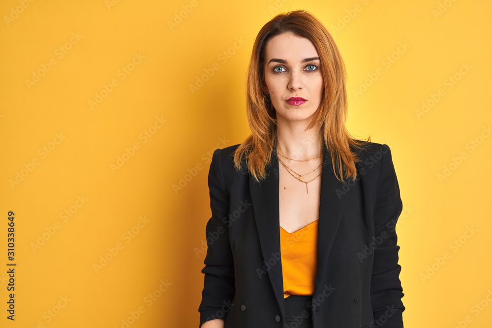 Redhead caucasian business woman over yellow isolated background with serious expression on face. Simple and natural looking at the camera. Redhead caucasian business woman over yellow isolated background with serious expression on face. Simple and natural looking at the camera.