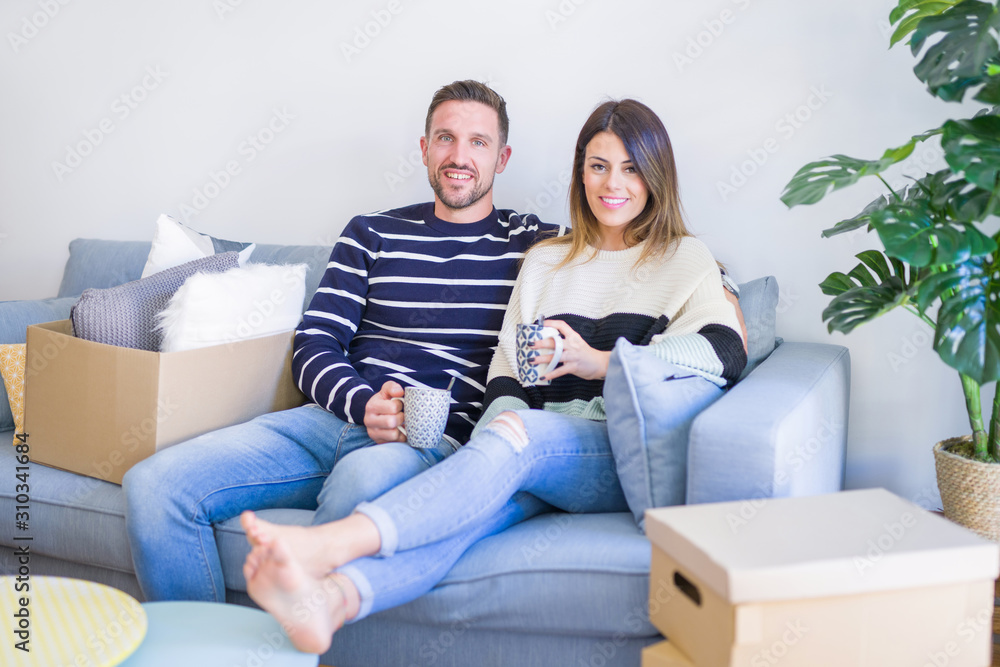 Young beautiful couple sitting on the sofa drinking cup of coffee at new home around cardboard boxes