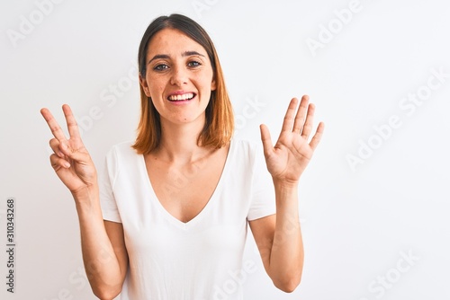 Beautiful redhead woman wearing casual white t-shirt over isolated background showing and pointing up with fingers number seven while smiling confident and happy.