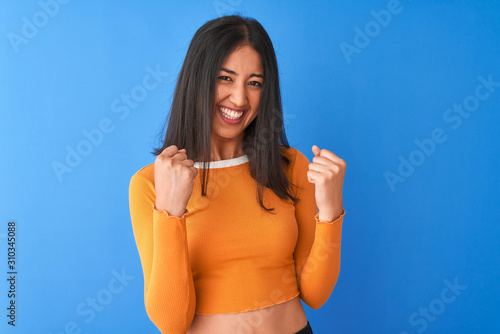 Obraz na plátně Young beautiful chinese woman wearing orange t-shirt standing over isolated blue background very happy and excited doing winner gesture with arms raised, smiling and screaming for success
