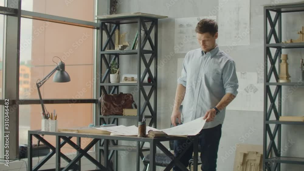 Engineer works in a bright office with a large window, concentrates and draws blueprints. Workplace of an architect or designer: loft style, minimalistic interior, drawings and coffee on the table Engineer works in a bright office with a large window, concentrates and draws blueprints. Workplace of an architect or designer: loft style, minimalistic interior, drawings and coffee on the table