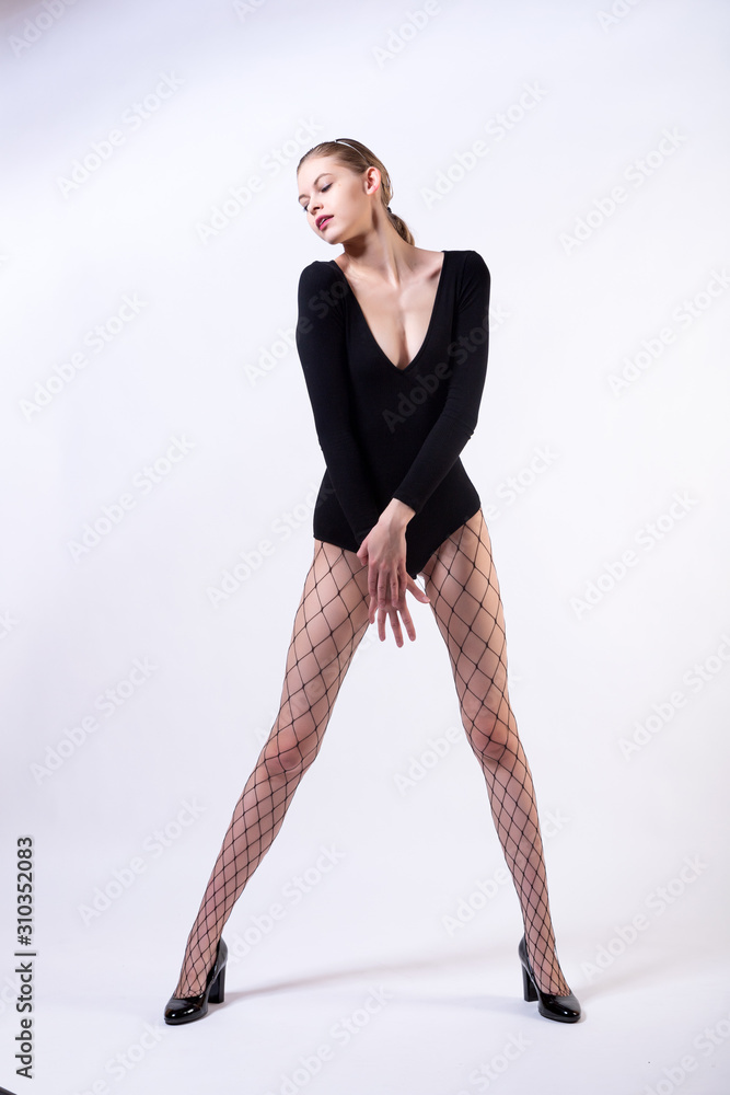 Young beautiful woman wearing in black body, fishnet tights and high heels posing over white background. Studio shot of redhaired woman against white background. Trendy fashionable Young beautiful woman wearing in black body, fishnet tights and high heels posing over white background. Studio shot of redhaired woman against white background. Trendy fashionable