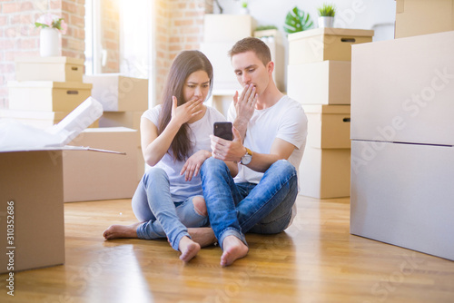 Beautiful couple sitting on the floor using smartphone at new home around cardboard boxes cover mouth with hand shocked with shame for mistake, expression of fear, scared in silence, secret concept © Krakenimages.com