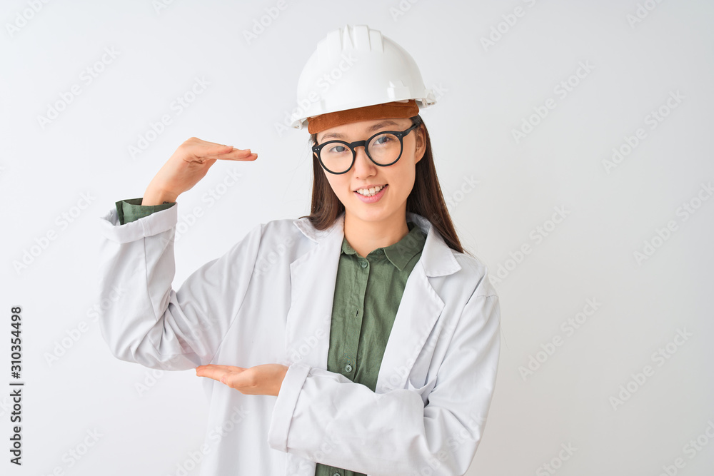 Young chinese engineer woman wearing coat helmet glasses over isolated white background gesturing with hands showing big and large size sign, measure symbol. Smiling looking at the camera. Measuring Young chinese engineer woman wearing coat helmet glasses over isolated white background gesturing with hands showing big and large size sign, measure symbol. Smiling looking at the camera. Measuring