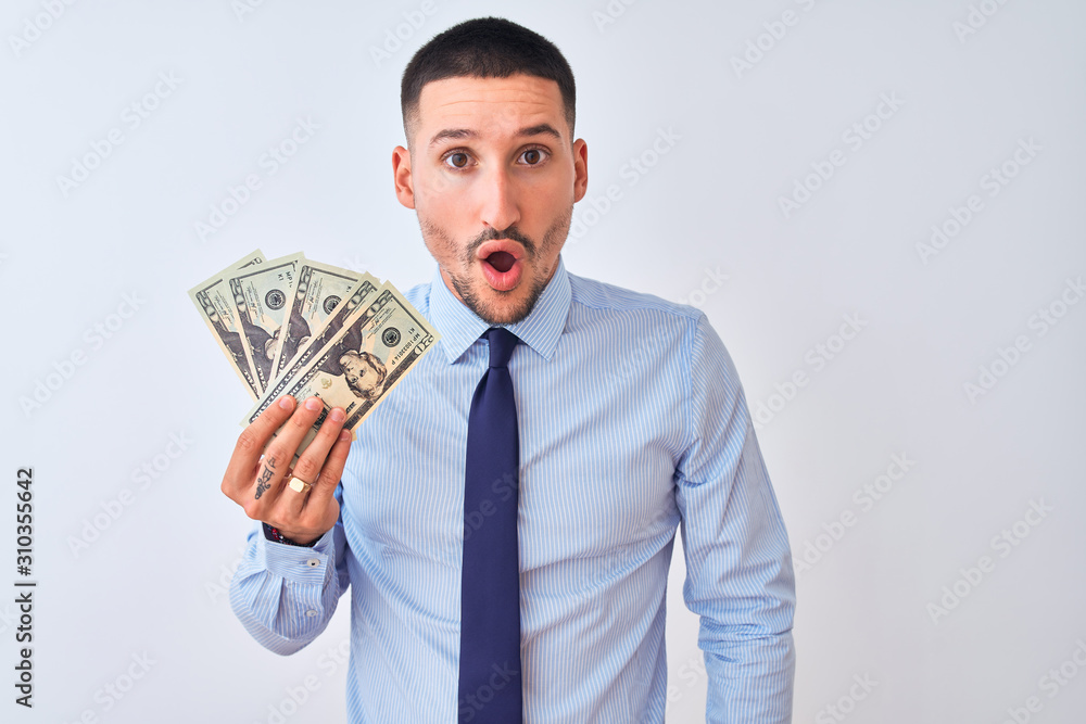 Young handsome business man holding a bunch of bank notes dollars over isolated background scared in shock with a surprise face, afraid and excited with fear expression Young handsome business man holding a bunch of bank notes dollars over isolated background scared in shock with a surprise face, afraid and excited with fear expression