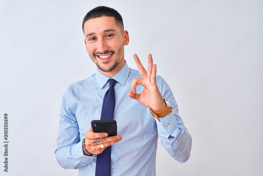 Young handsome business man using smartphone over isolated background doing ok sign with fingers, excellent symbol Young handsome business man using smartphone over isolated background doing ok sign with fingers, excellent symbol