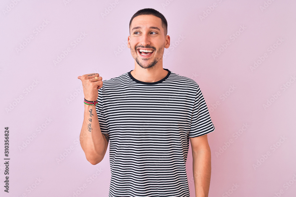 Young handsome man wearing striped t-shirt standing over pink isolated background smiling with happy face looking and pointing to the side with thumb up. Young handsome man wearing striped t-shirt standing over pink isolated background smiling with happy face looking and pointing to the side with thumb up.