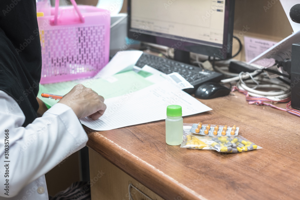 Thai young muslim doctor pharmacist wearing hijab in uniform write medical prescription at desk with drug pills and a computer. Thai young muslim doctor pharmacist wearing hijab in uniform write medical prescription at desk with drug pills and a computer.