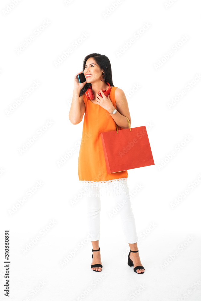 A beautiful Asian young woman wearing orange dress holding a red shopping bag talking on mobile phone with red head phone around her neck in front of white isolated background. A beautiful Asian young woman wearing orange dress holding a red shopping bag talking on mobile phone with red head phone around her neck in front of white isolated background.