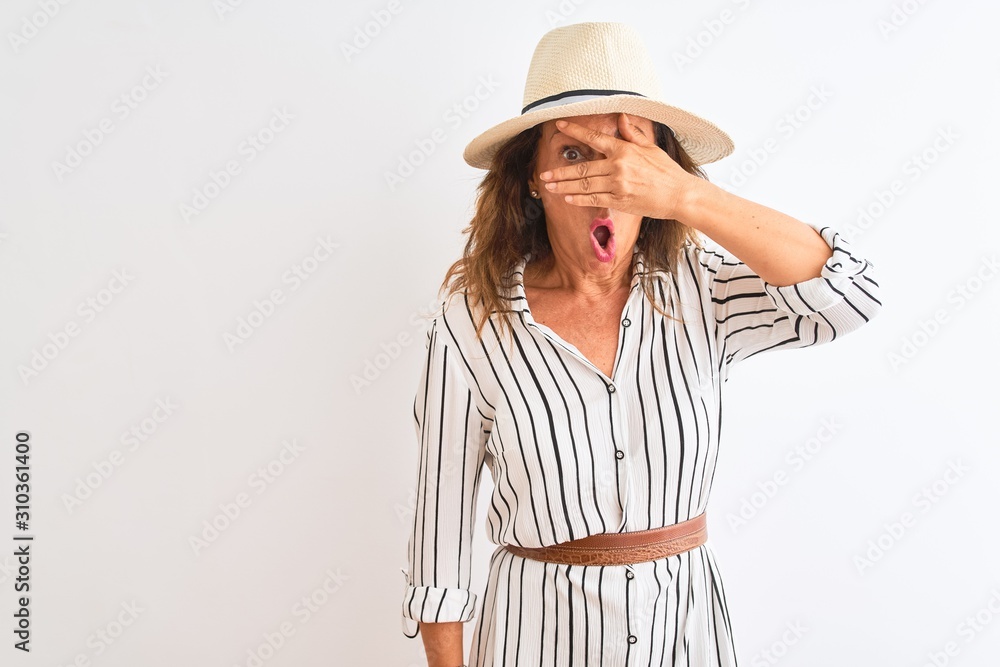Middle age businesswoman wearing striped dress and hat over isolated white background peeking in shock covering face and eyes with hand, looking through fingers with embarrassed expression. Middle age businesswoman wearing striped dress and hat over isolated white background peeking in shock covering face and eyes with hand, looking through fingers with embarrassed expression.