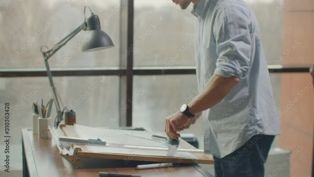 A man is a young modern architect sitting at a table drawing a plan of the building and developing a design, sitting in an office in the Sitel loft near a large window. A man is a young modern architect sitting at a table drawing a plan of the building and developing a design, sitting in an office in the Sitel loft near a large window.