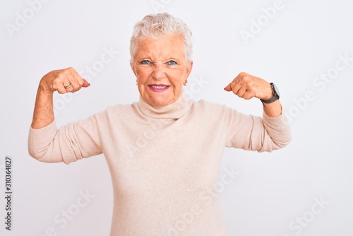 Senior grey-haired woman wearing turtleneck sweater standing over isolated white background showing arms muscles smiling proud. Fitness concept.