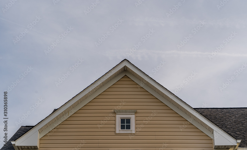 Single gable close up with beige vinyl siding on luxury single family residential home with single attic window in white frame with neutral sky background