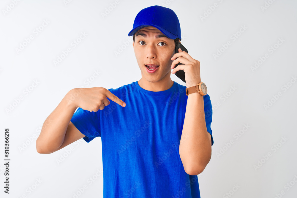 Chinese deliveryman wearing cap talking on the smartphone over isolated white background with surprise face pointing finger to himself