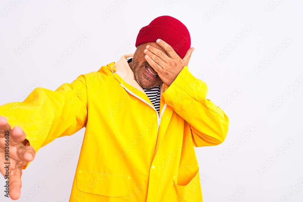 Middle age man wearing rain coat and woolen hat standing over isolated white background covering eyes with hands and doing stop gesture with sad and fear expression. Embarrassed and negative concept.