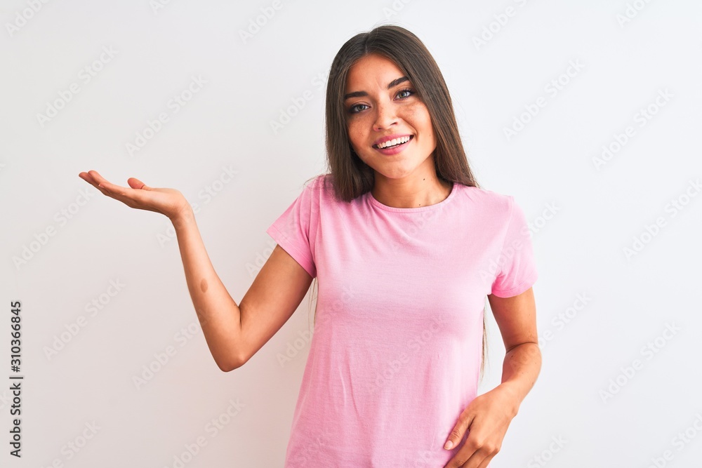Young beautiful woman wearing pink casual t-shirt standing over isolated white background smiling cheerful presenting and pointing with palm of hand looking at the camera. Young beautiful woman wearing pink casual t-shirt standing over isolated white background smiling cheerful presenting and pointing with palm of hand looking at the camera.