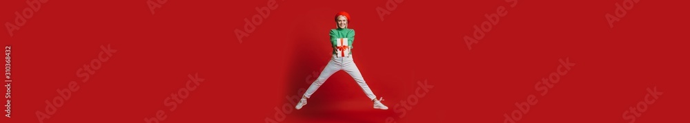 Full length of a lovely woman holding a red gift box jumping laughing against a red background.