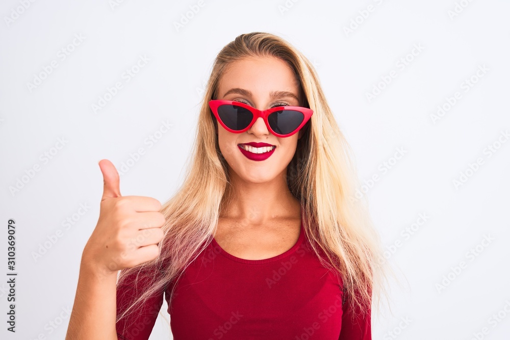 Young beautiful woman wearing red t-shirt and sunglasses over isolated white background doing happy thumbs up gesture with hand. Approving expression looking at the camera with showing success. Young beautiful woman wearing red t-shirt and sunglasses over isolated white background doing happy thumbs up gesture with hand. Approving expression looking at the camera with showing success.