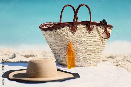 Women's beach accessories on sand for summer vacation concept. Straw tote bag, sun hat and sunscreen lotion or suntan tanning oil spray bottle with blue ocean background for travel holidays.