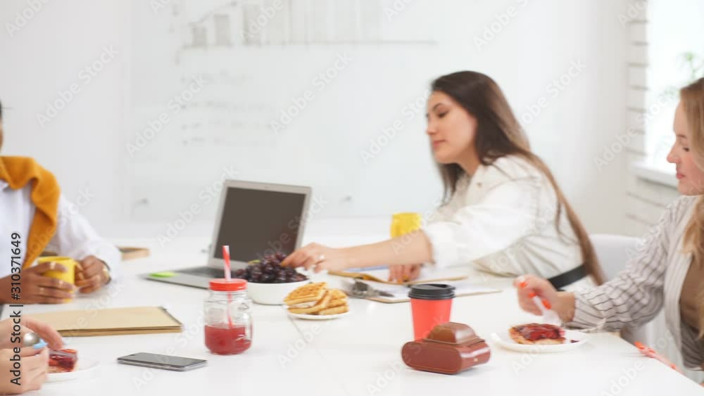 Young four women sitting on table in office, drinking and eating suring break at workplace Young four women sitting on table in office, drinking and eating suring break at workplace