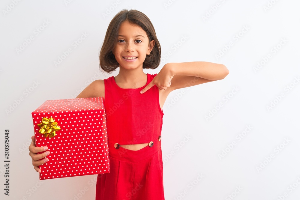 Beautiful child girl holding birthday gift standing over isolated white background with surprise face pointing finger to himself