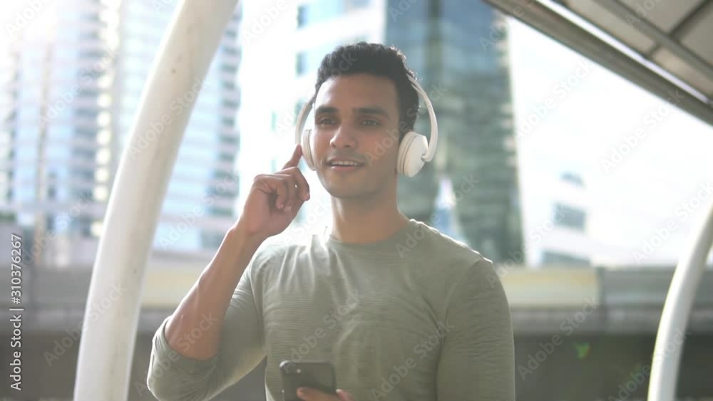 Portrait of a smiling young Indian man holding mobile phone in hand enjoying listening to music on wireless headphone Portrait of a smiling young Indian man holding mobile phone in hand enjoying listening to music on wireless headphone