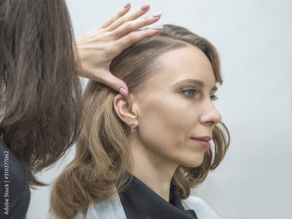 Fototapeta premium Women make-up artist and hairdresser do hairstyle and stylish makeup for beautiful middle age blonde girl before start of fashion show. Colorful makeup design beauty and art salon concept.