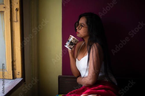 An young and attractive Indian brunette woman in white sleeping wear with a coffee/tea cup reading book on a bed inside in her room. Indian lifestyle.