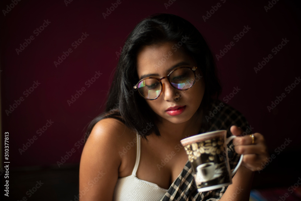 n young and attractive Indian Bengali brunette woman in white sleeping wear and spectacles with a coffee/tea cup on a bed inside in her room. Indian lifestyle. n young and attractive Indian Bengali brunette woman in white sleeping wear and spectacles with a coffee/tea cup on a bed inside in her room. Indian lifestyle.