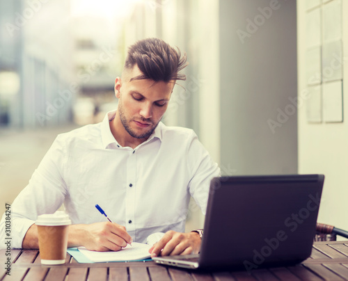 business, education, technology and people concept - young man with laptop computer, documents and coffee cup at city street cafe