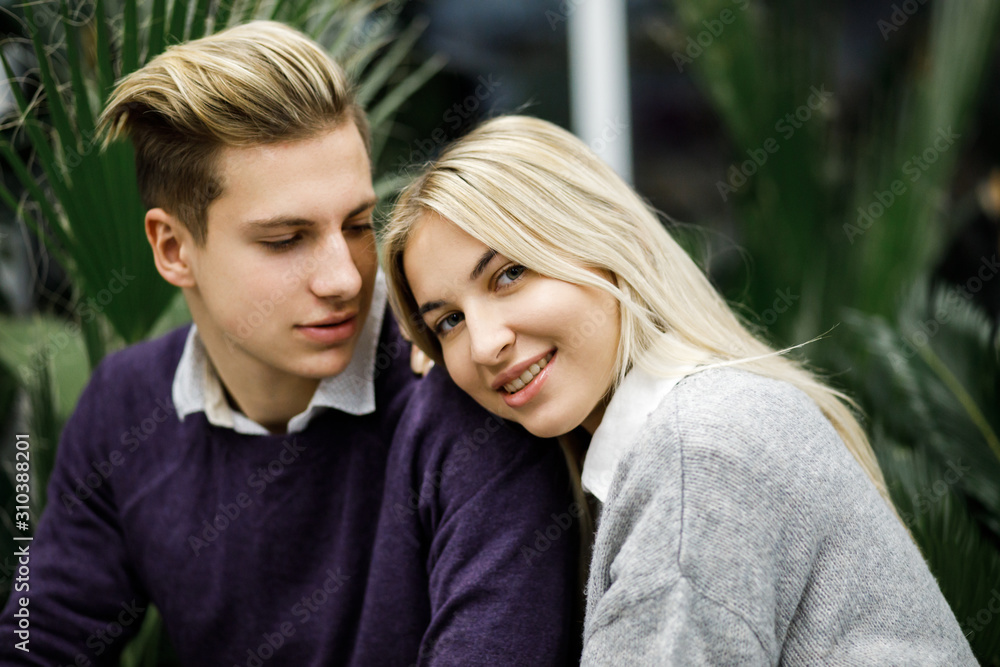 Cute blond guy and girl sitting among tropical plants in big store. Shop for decor Cute blond guy and girl sitting among tropical plants in big store. Shop for decor