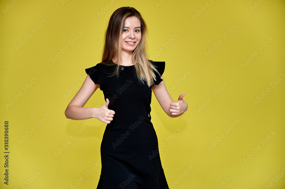 Portrait of a pretty young smiling woman on a yellow background in a black dress with long straight hair. Standing right in front of the camera, Shows emotions, talks in different poses. Portrait of a pretty young smiling woman on a yellow background in a black dress with long straight hair. Standing right in front of the camera, Shows emotions, talks in different poses.